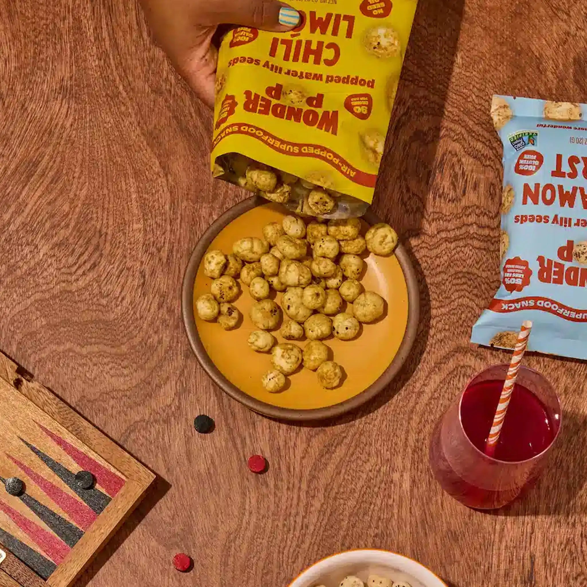 Hand pouring pops into a bowl on a wooden table with a drink and game board.